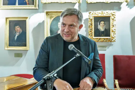 Mężczyzna przemawia przy mikrofonie, w tle portrety na ścianie.