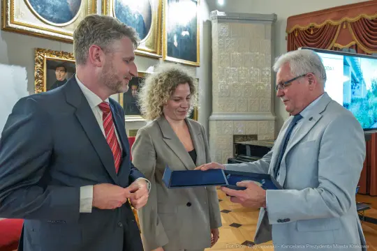 Wręczenie nagrody przez przedstawiciela miasta dwóm osobom w eleganckim wnętrzu.