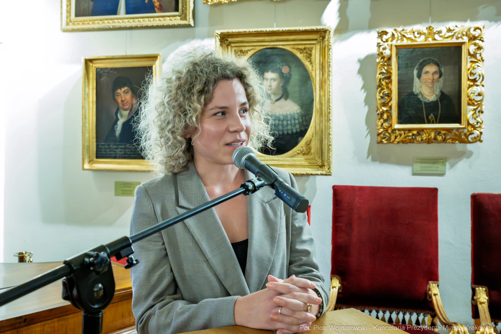 Kobieta z kręconymi włosami przemawia do mikrofonu w otoczeniu portretów.  Autor: Piotr Wojnarowski