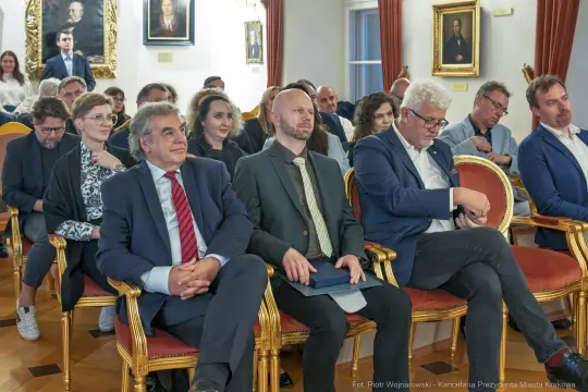 Grupa ludzi siedzących na eleganckich krzesłach w sali z portretami na ścianach.