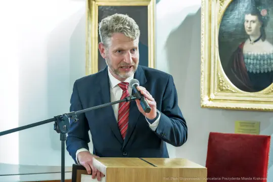 Mężczyzna w garniturze przemawia do mikrofonu w eleganckim wnętrzu.