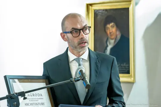 Mężczyzna w garniturze trzyma nagrodę i przemawia do mikrofonu. W tle portret.