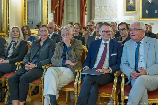 Grupa osób siedzących w eleganckiej sali, uczestniczących w wydarzeniu.