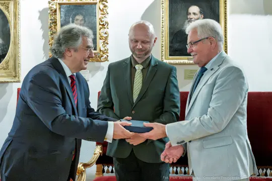 Trzech mężczyzn wymienia się prezentami w eleganckim wnętrzu z portretami na ścianach.