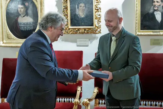 Dwaj mężczyźni wymieniają się książką w eleganckim otoczeniu z portretami na ścianach.