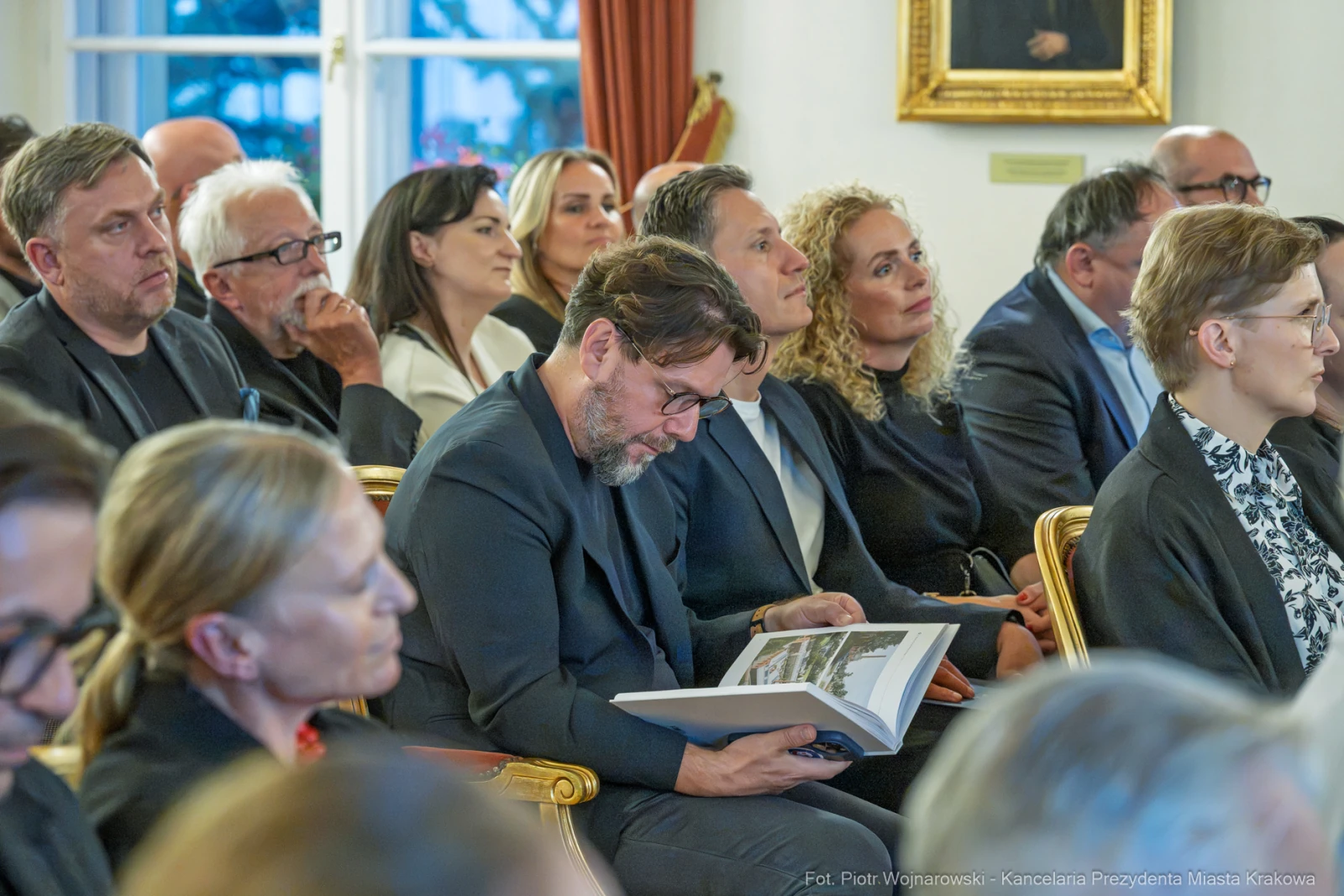 Grupa ludzi w eleganckich strojach, niektórzy siedzą, inni przeglądają książkę.  Autor: Piotr Wojnarowski