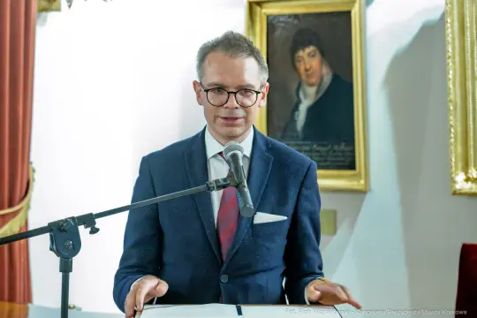Mężczyzna w garniturze przemawia przy mównicy, w tle portret.