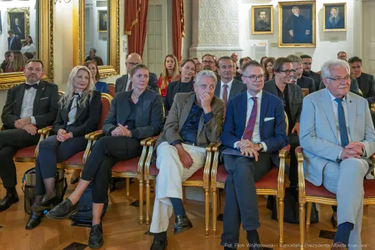 Grupa osób siedzących w eleganckiej sali, w tle portrety na ścianach.