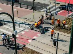 Prace drogowe w mieście, robotnicy w pomarańczowych kamizelkach, ruch pieszych i rowerzystów.