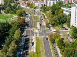 Widok na ulicę z torami tramwajowymi, zieleń i zabudowę miejską w tle.