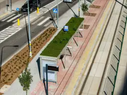 Nowoczesny przystanek tramwajowy z zielonym dachem, otoczony roślinnością i ulicą.