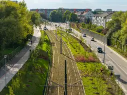Zielona przestrzeń z torami tramwajowymi, ścieżkami dla pieszych i rowerów oraz zabudową mieszkalną w tle.