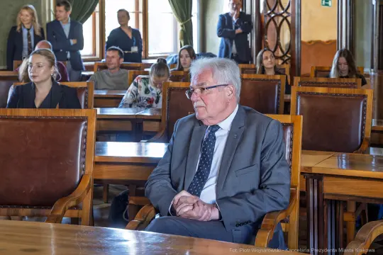 Mężczyzna w garniturze siedzi w sali z innymi osobami, w tle drewniane meble i okna.