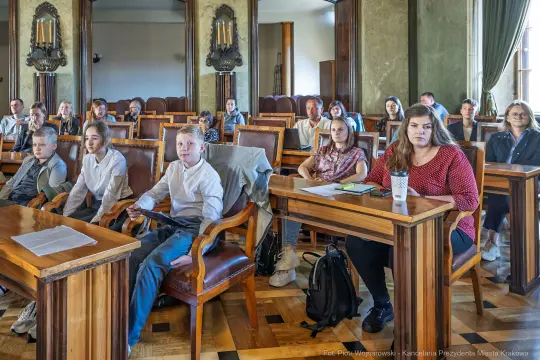 Grupa dzieci i dorosłych siedzących w sali, uczestniczących w spotkaniu.