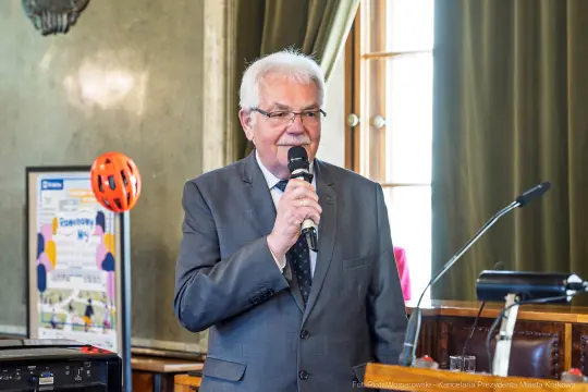Mężczyzna w garniturze przemawia do mikrofonu w eleganckim wnętrzu.