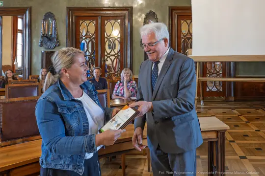 Wręczenie nagrody przez mężczyznę kobiecie w sali z drewnianymi meblami.