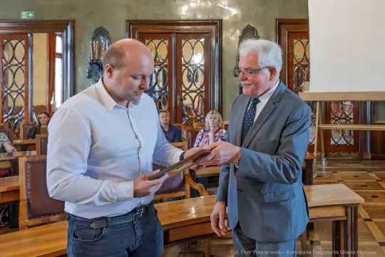 Mężczyzna odbierający nagrodę od drugiego mężczyzny w sali z drewnianymi elementami.