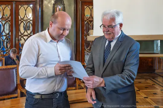 Dwaj mężczyźni wymieniają się dokumentami w eleganckim wnętrzu.