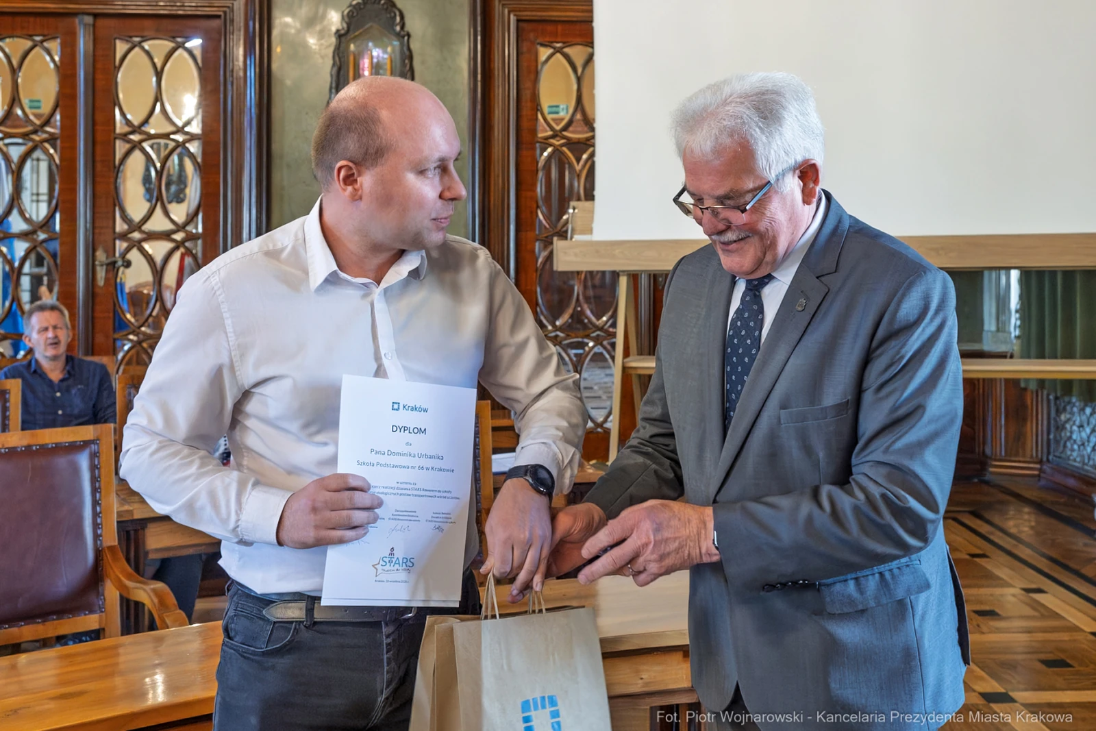 Mężczyzna odbierający dyplom od innego mężczyzny w eleganckim wnętrzu.  Autor: Piotr Wojnarowski