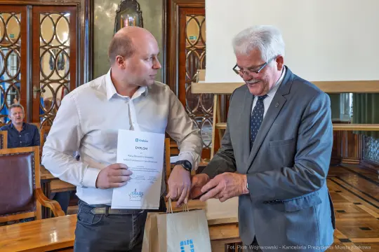 Mężczyzna odbierający dyplom od innego mężczyzny w eleganckim wnętrzu.