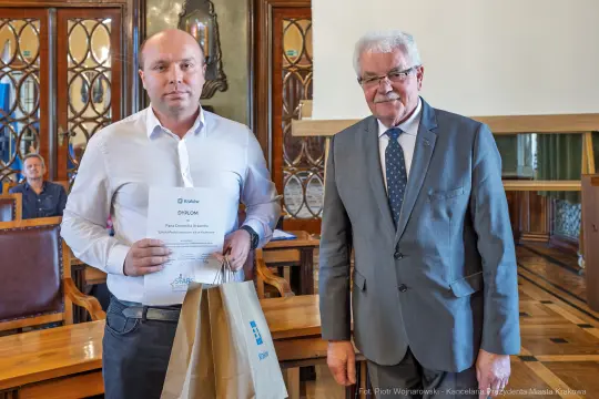 Mężczyzna trzyma dyplom i torbę, obok stoi starszy mężczyzna w garniturze.