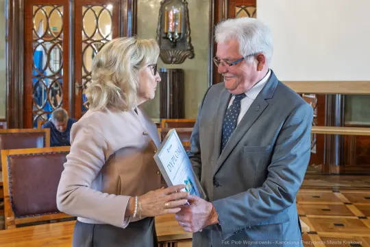 Wręczenie nagrody w eleganckim wnętrzu, uśmiechnięte osoby wymieniają się dokumentem.