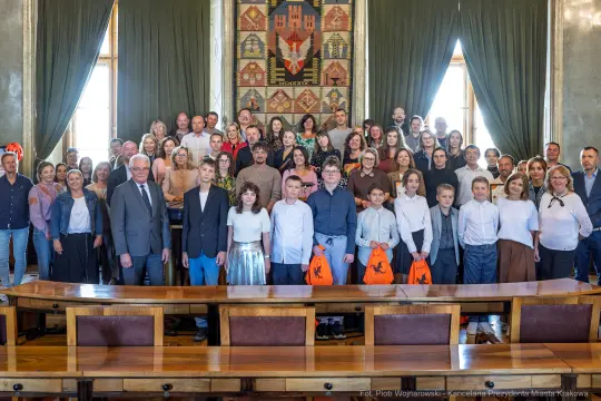Grupa ludzi w sali, ubranych w eleganckie stroje, z torbami w rękach.