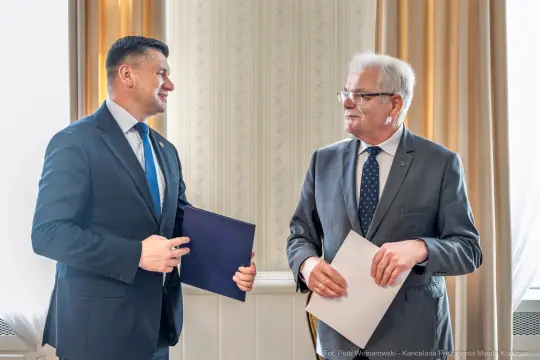 Dwaj mężczyźni w garniturach, rozmawiający i trzymający dokumenty.