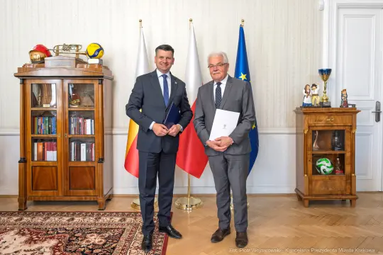 Dwaj mężczyźni w formalnych strojach, stojący obok siebie w biurze z flagami i meblami.