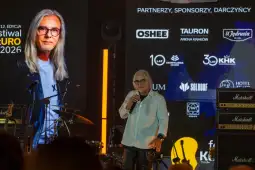 Mężczyzna z okularami i długimi włosami przemawia na scenie festiwalu muzycznego.