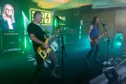 Zespół grający na żywo, z gitarzystami i perkusistą, w tle kolorowe światła i sprzęt muzyczny.