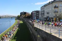 Biegaczy na trasie wzdłuż rzeki, otoczeni budynkami i zielenią.