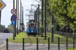 Tramwaj na torach wzdłuż drogi, otoczony zielenią i znakami drogowymi.