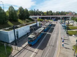 Tramwaje na przystanku, pasażerowie czekający na transport, zieleń w tle.
