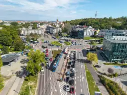 Ruchliwa ulica z tramwajem i samochodami, w tle zabudowa miejska i zieleń.