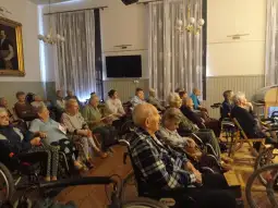Grupa starszych osób siedzących w sali, część z nich na wózkach inwalidzkich.