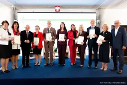 Grupa osób trzymających dokumenty, na tle sali konferencyjnej z herbem Polski.