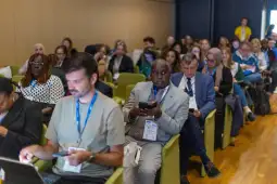 Grupa ludzi siedzących w sali konferencyjnej, niektórzy korzystają z telefonów i laptopów.