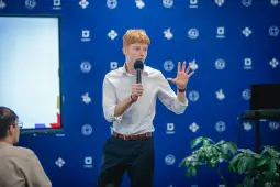 Mężczyzna w białej koszuli przemawia do publiczności, trzymając mikrofon.