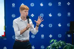 Młody mężczyzna w białej koszuli przemawia do publiczności, trzymając mikrofon.