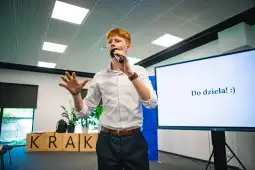 Młody mężczyzna przemawia do publiczności, trzymając mikrofon, z napisem "Do dzieła!" na ekranie.