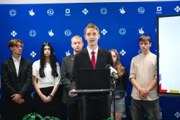 Młody mężczyzna przemawia z mównicy, za nim grupa ludzi w formalnych strojach.