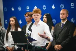 Młody mężczyzna przemawia na konferencji, w tle grupa osób i niebieska ściana z logo.