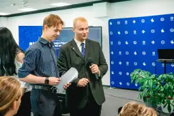 Dwóch mężczyzn na scenie, jeden z mikrofonem, drugi trzyma kartkę. Tło z logo i grafiką.