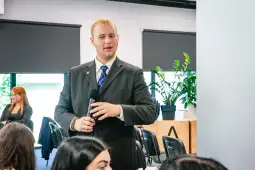 Mężczyzna w garniturze trzyma mikrofon, prowadzi dyskusję w sali konferencyjnej.