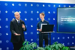 Dwóch mężczyzn podczas prezentacji, jeden z mikrofonem, drugi z laptopem. Tło z logo.