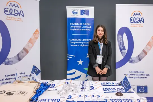 Kobieta stoi przy stoisku z materiałami promującymi lokalną demokrację.