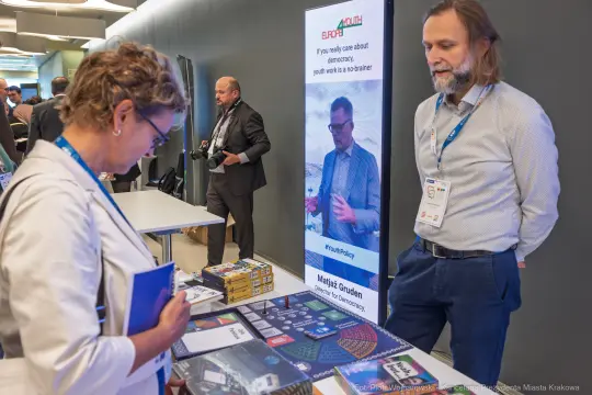 Kobieta przegląda materiały promocyjne na stoisku podczas konferencji. W tle mężczyzna rozmawia z innymi uczestnikami.