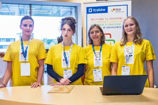Cztery osoby w żółtych koszulkach stoją przy biurku, promując Kraków jako miejsce wydarzenia.
