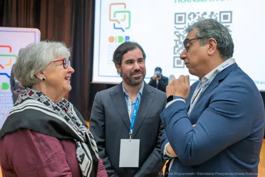 Trzy osoby rozmawiające podczas konferencji, w tle ekran z kodem QR.